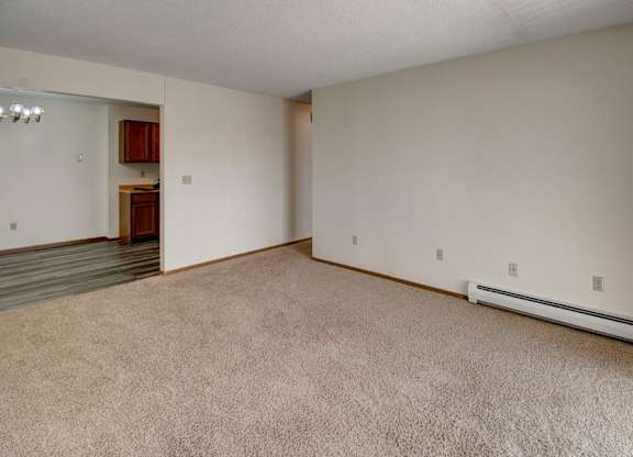 A spacious, open living area here at Windscape Apartments with soft neutral carpeting, light-colored walls, and clean baseboard trim. The room offers flexible layout options with multiple outlets along the wall and a clear connection to the adjacent dining and kitchen area, creating a bright and versatile space for everyday living.