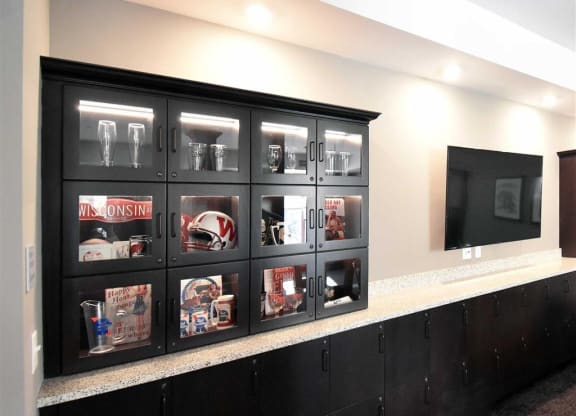 Black display cabinet with sports memorabilia and glasses at the Oaks of Shorewood Apartments, set against a light wall next to a large flat-screen TV. Modern and organized ambiance.