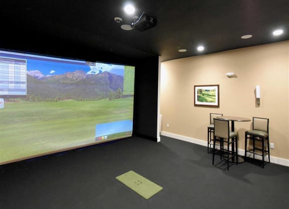 Indoor golf simulator at the Oaks of Shorewood Apartments with a large screen displaying a golf course scene. To the right, a small table with chairs and a framed picture on the wall.