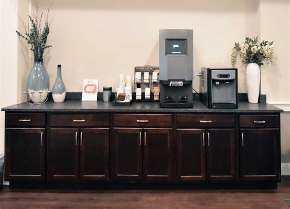 Coffee station with a modern coffee machine and water dispenser on a dark wooden cabinet at the Oaks of Shorewood Apartments. Decorated with tall vases and flowers, creating a welcoming atmosphere.