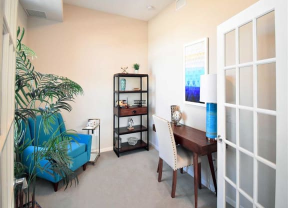 Cozy office at the Oaks of Shorewood Apartments with a blue armchair, potted plant, and shelf against cream walls. A wooden desk has a white chair, lamp, and abstract blue artwork.