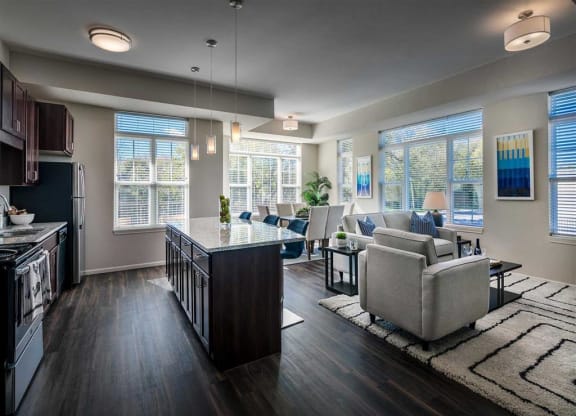 Modern open-concept kitchen and living room with dark wood floors and sleek furniture. Large windows let in natural light, creating a cozy, inviting atmosphere at the Oaks of Shorewood Apartments.