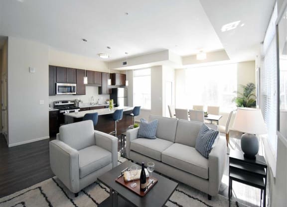 Modern open-concept living area at the Oaks of Shorewood Apartments with a cozy gray sofa, armchair, and coffee table. Bright kitchen and dining space in the background with natural light.
