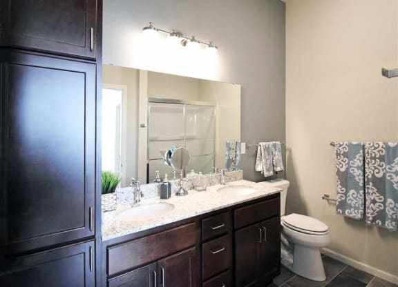 Modern bathroom at the Oaks of Shorewood Apartments with dark wood cabinets, dual sinks, and granite countertop. Wall-mounted lights illuminate the space. Stylish towels hang near the toilet.