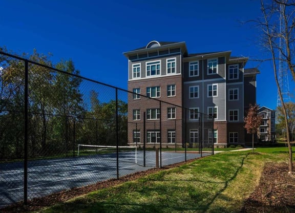 Four-story brick building with white-trimmed windows beside a fenced tennis court. Surrounded by trees under a clear blue sky, creating a peaceful atmosphere. The Oaks of Shorewood Apartments.