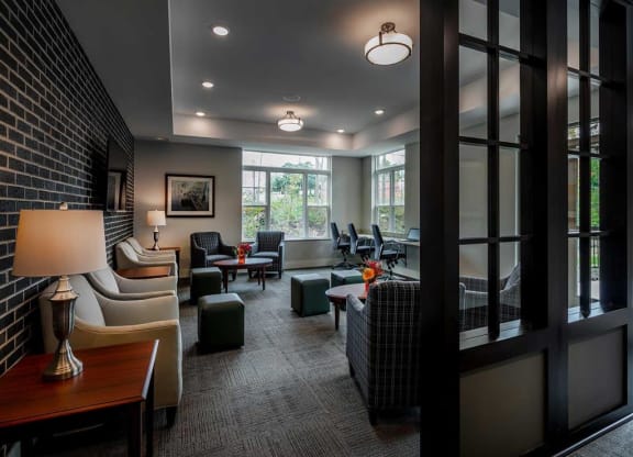 Modern lounge at the Oaks of Shorewood Apartments with soft lighting features beige armchairs, plaid chairs, and dark wood accents. Large windows provide natural light, creating a cozy atmosphere.