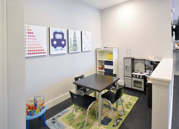 Bright playroom with a small table and four chairs on a colorful rug. Wall art shows numbers, a car, buildings, and the alphabet. Toy shelves and a pretend kitchen set give a playful vibe at the Oaks of Shorewood Apartments.