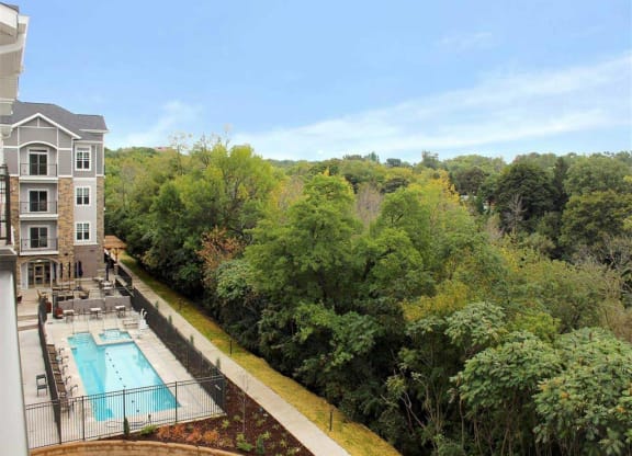 Oaks of Shorewood, a modern apartment building beside a pool with lounge chairs, bordered by lush green trees under a clear blue sky. Calm and inviting outdoor setting.