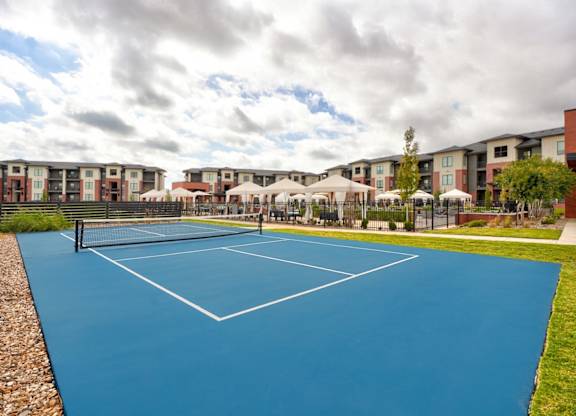 A blue tennis court with white lines is surrounded by a gravel area and a grassy area.