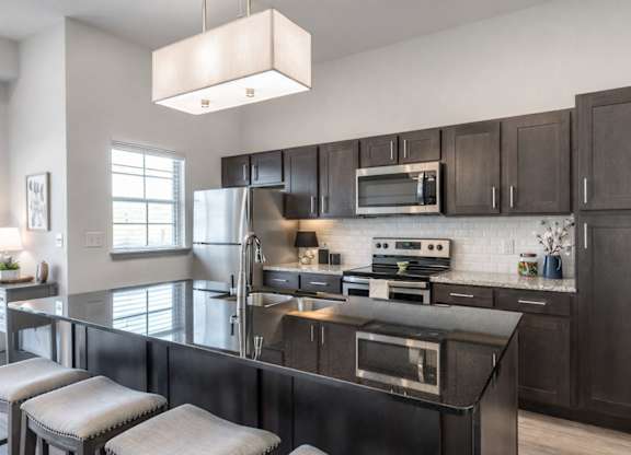 A sleek kitchen here at 98 Apartments with dark cabinetry, stainless steel appliances, glossy granite countertops, a large island with seating, and bright lighting, creating a modern and polished space for cooking and entertaining.