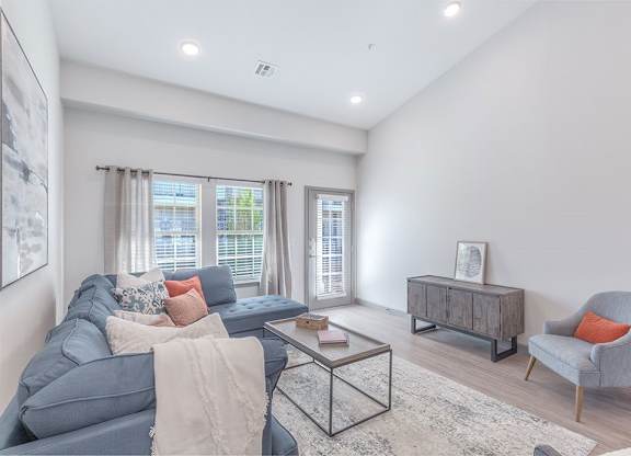A bright living room here at 98 Apartments with wood-style flooring, a comfortable sectional sofa, soft neutral décor, and large windows with a glass door that bring in natural light, creating a calm and inviting space for everyday living.