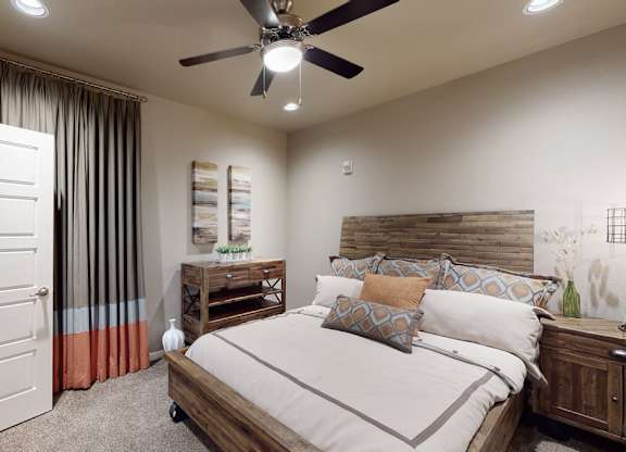 A warm bedroom here at 98 Apartments with soft carpet, a wood-accent bed frame, neutral bedding with patterned pillows, a ceiling fan, and floor-length curtains, creating a cozy and restful retreat.