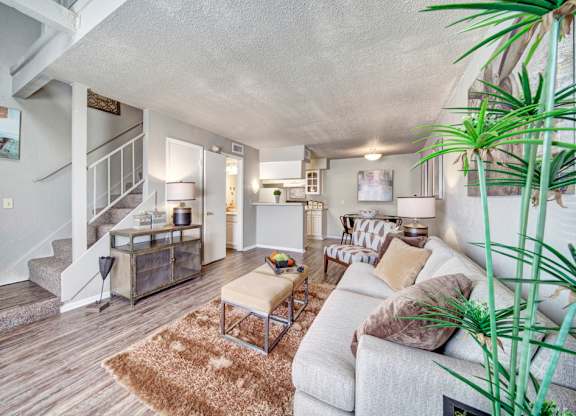 A cozy living room here at Brookwood Village with wood-style flooring, soft neutral furnishings, and warm lighting, featuring an open layout with a staircase, a small dining area, and a connected kitchen that creates an inviting and functional living space.