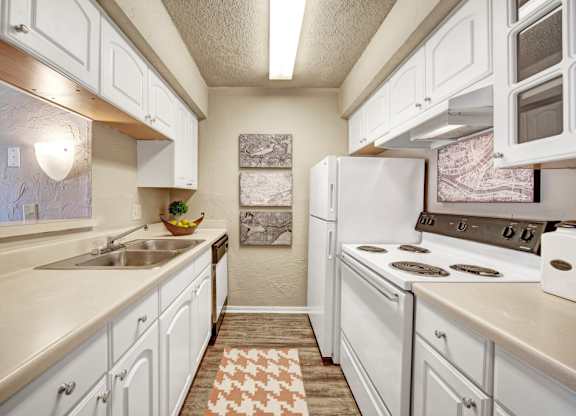 A bright kitchen here at Brookwood Village with white cabinets, light countertops, and white appliances, featuring a galley-style layout, wood-style flooring, and soft neutral accents that create a clean and welcoming cooking space.
