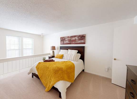 A bright bedroom here at Brookwood Village with beige carpet, soft neutral walls, and natural light from multiple windows, featuring a cozy bed with warm accent bedding and an open, airy layout that feels calm and inviting.