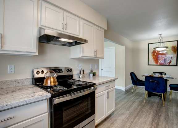 A bright kitchen here at Indigo with white cabinetry, light countertops, and black appliances including an oven and range hood. A nearby dining area with modern lighting and comfortable seating creates a clean, inviting space for everyday living.