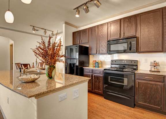 Here at Liberty Pointe, a bright kitchen with rich dark-wood cabinetry, black appliances, and warm wood-tone flooring. The granite breakfast bar features a decorative bowl and a tall greenery arrangement centerpiece, with track and pendant lighting above and a view into the adjacent dining area.