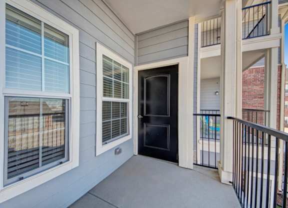 A private exterior entry balcony here at Liberty Pointe featuring gray siding, white trim windows, and a black front door. The space includes metal railing details and a covered walkway, creating a clean and welcoming exterior approach.