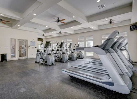 A fitness center here at Liberty Pointe featuring treadmills, stationary bikes, and elliptical machines arranged across an open workout floor. The space has bright recessed lighting, ceiling fans, and multiple windows that bring in natural light for a clean, energized atmosphere.