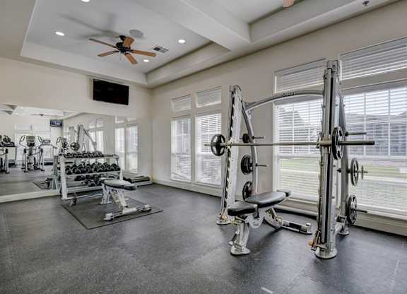 A fitness center here at Liberty Pointe featuring a weight training area with a squat rack, adjustable bench, and free weights. Large windows and mirrors create a bright, open workout space with a clean and modern feel.