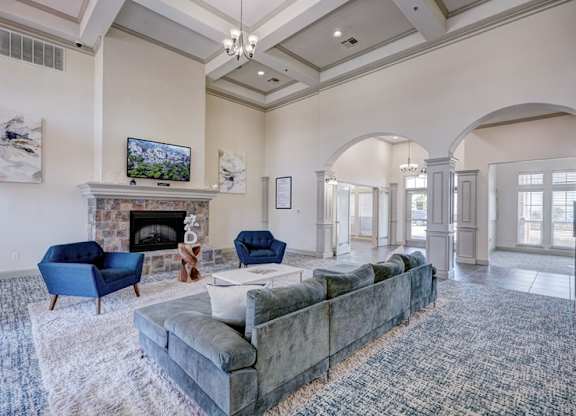 A resident lounge here at Liberty Pointe featuring a plush sofa and blue accent chairs arranged around a coffee table, with a stone fireplace and mounted TV as the focal point. Tall ceilings, arched architectural details, and soft natural light create a bright, comfortable space for residents to gather and relax.