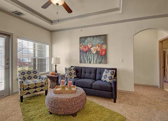 Here at Liberty Pointe, a cozy living area with a deep blue sofa and a patterned accent chair paired with a bold green rug. A woven round ottoman coffee table sits centered with tray décor and candles, while a tulip wall art piece adds warm color above the sofa and natural light comes through the window and patio door.