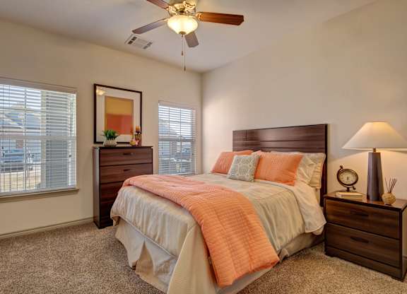 Here at Liberty Pointe, a bright bedroom with warm neutral walls and soft carpet, featuring a wooden bed frame styled with a white bedspread and standout coral bedding accents. Matching wood nightstand and dresser add warmth, while a table lamp, framed wall art, and natural light from two windows create a clean, comfortable feel.