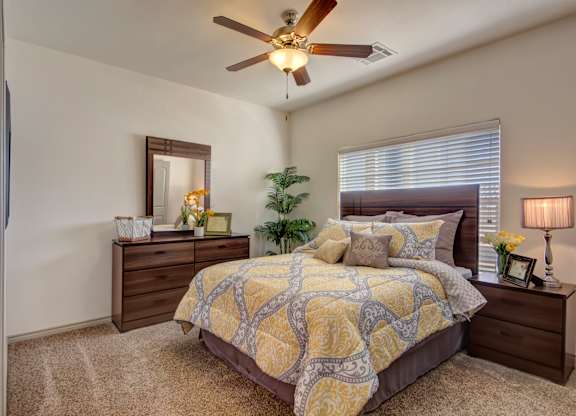 A bedroom here at Liberty Pointe featuring soft neutral walls, plush carpet, and a bed with yellow and gray patterned bedding and layered neutral accent pillows. The space includes dark wood nightstands and a dresser. Includes a soft green plant in the corner of the space with a wooden ceiling fan and fan light, creating a cozy atmosphere.