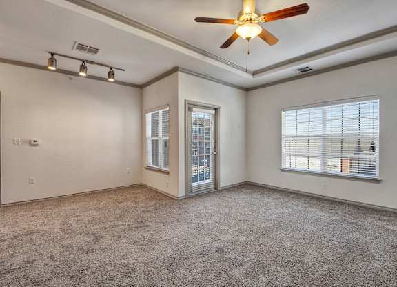 A living room here at Liberty Pointe featuring soft beige walls, plush neutral carpet, and bright natural light from multiple windows and a glass door. The space includes tray ceiling detail with a ceiling fan and modern track lighting, creating a bright, open, and comfortable atmosphere.