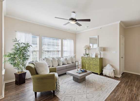 A living room here at Lincoln at Central Park featuring a bright, modern space with a light gray sofa, a green accent chair, and a green decorative dresser that adds a standout pop of color. Large windows with white curtains bring in natural light, while a light area rug, square coffee table, and indoor plant create a clean, comfortable feel.