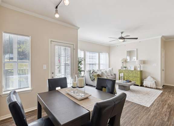 A dining and living area here at Lincoln at Central Park featuring a dark wood dining table with four black upholstered chairs, styled with a neutral table runner and glass candle centerpieces. The space opens into a bright living room with a light neutral sofa, a green accent chair, and a standout green decorative dresser that adds a fresh pop of color. Large windows bring in natural light.