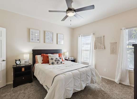 A bedroom here at Lincoln at Central Park featuring a dark wood bed frame styled with white bedding and standout rust-orange accent pillows and throw for a warm pop of color. Matching dark wood nightstands with modern lamps and décor sit on each side, while neutral wall art above the bed and soft white curtains over the windows keep the space bright, clean, and inviting.