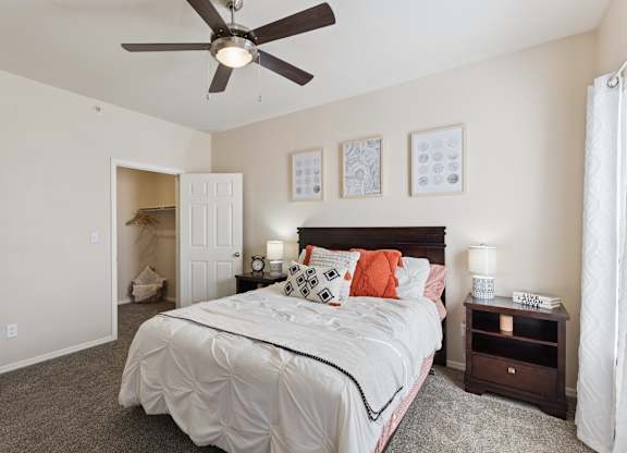 A bedroom here at Lincoln at Central Park featuring a dark wood bed, white bedding, and orange accent pillows. Includes matching nightstands, soft carpet, ceiling fan, and an open closet, creating a bright, cozy, and inviting space.