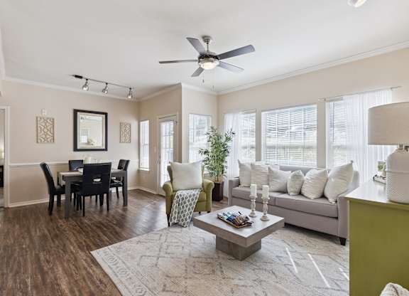 A living room here at Lincoln at Central Park featuring an open, bright layout with a light gray sofa, a green accent chair, and a green decorative dresser that adds a standout pop of color. Large windows with white curtains bring in natural light, while a light area rug, square coffee table, and nearby dining area create a comfortable, modern feel.