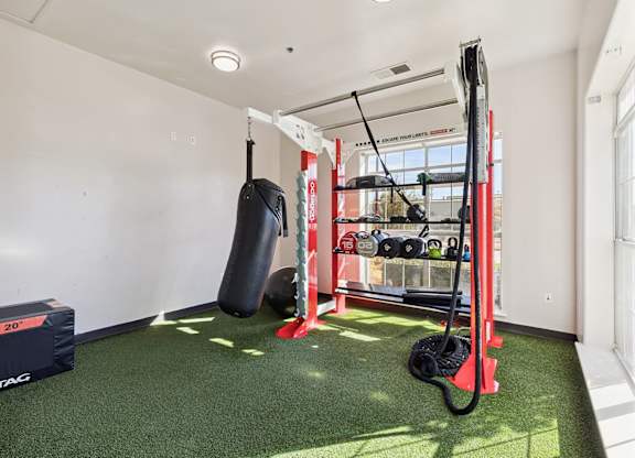 A fitness room here at Lincoln at Central Park with green turf flooring, a modern red workout rack, punching bag, kettlebells, and battle ropes. A large window brings in bright natural light, creating an energetic, clean workout environment.
