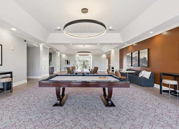 A modern resident lounge here at Lincoln at Central Park with plush carpet flooring, a pool table centerpiece, and stylish circular ceiling lighting. A warm accent wall, comfortable seating, and dining space create a welcoming, upscale social atmosphere.