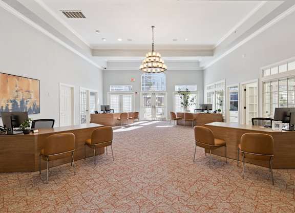 A welcoming leasing office lobby here at Lincoln at Central Park with wood desks, comfortable guest seating, and soft patterned carpet. A large chandelier and tall windows create a bright, upscale, and professional first impression for residents and visitors.