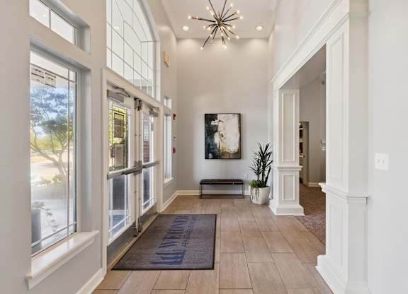 A bright entry lobby here at Lincoln at Central Park with tall windows, glass entry doors, and light wood-style flooring. A modern chandelier, decorative artwork, and greenery create a clean, welcoming first impression for residents and guests.