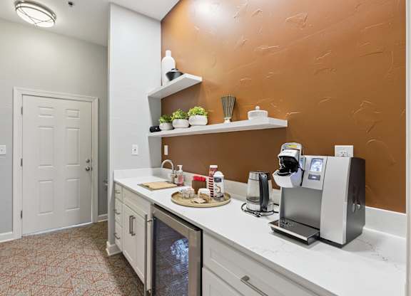 A resident coffee station here at Lincoln at Central Park with white cabinetry, quartz-style countertops, and a modern coffee machine. A mini fridge, sink, and decorative shelving create a convenient, stylish refreshment area for residents.