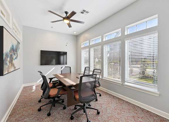 A private meeting room here at Lincoln at Central Park with a wood conference table, rolling office chairs, and a wall-mounted TV. A ceiling fan and large windows with blinds create a bright, comfortable workspace for meetings or remote work.