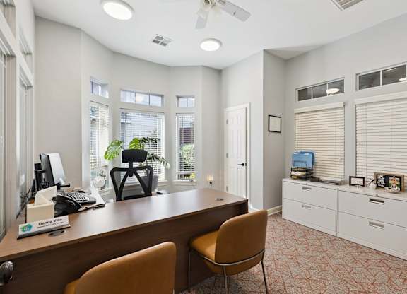 A private office here at Lincoln at Central Park with a wood desk, guest seating, and built-in file storage. A ceiling fan and multiple windows with blinds create a bright, comfortable workspace with natural light.