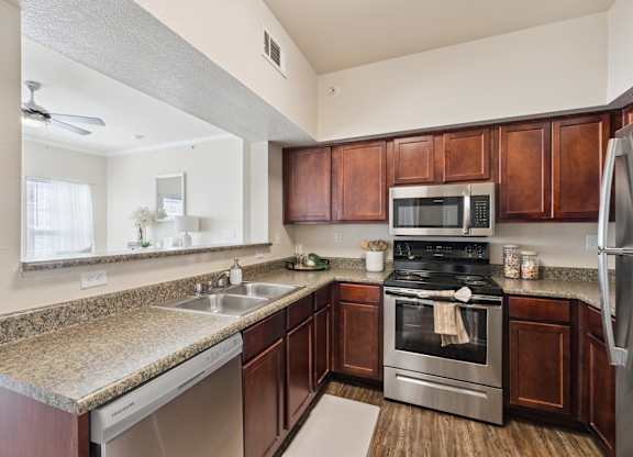 A kitchen here at Lincoln at Central Park featuring rich wood cabinetry, granite-style countertops, and stainless steel appliances including a microwave, range, dishwasher, and refrigerator. The space includes a double-basin sink with a pass-through opening to the living area, simple countertop accessories, and wood-style flooring, creating a bright and clean cooking space.
