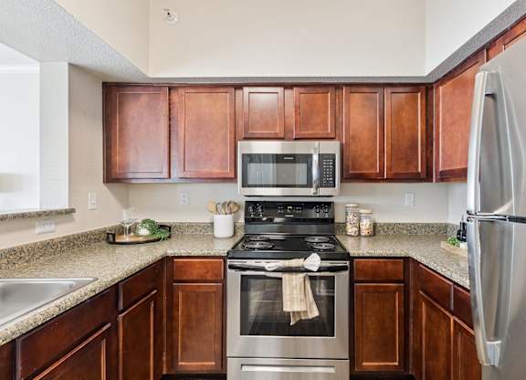 A kitchen here at Lincoln at Central Park featuring warm wood cabinetry, granite-style countertops, and stainless steel appliances including a microwave, range, and refrigerator. The space includes simple countertop décor like glassware, jars, and a small tray with greenery, paired with neutral walls and wood-style flooring for a bright, clean, and inviting cooking area.