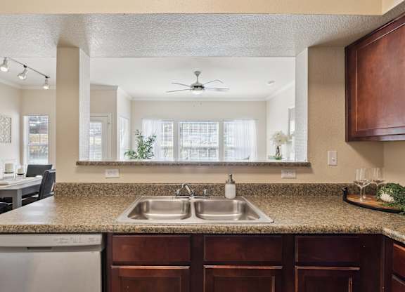 A kitchen here at Lincoln at Central Park featuring a double stainless steel sink set into granite-style countertops with rich wood cabinetry below. The pass-through bar opens to a bright living area with large windows and sheer curtains, while simple accents like a soap dispenser, glassware, and a small greenery tray create a clean, modern, and welcoming space.