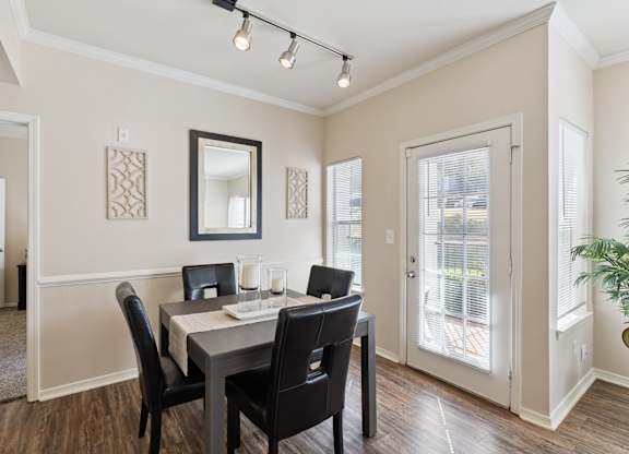 A dining area here at Lincoln at Central Park featuring a dark wood dining table paired with four black upholstered chairs, set on warm wood-style flooring. Natural light enters through a glass patio door and side window with white blinds, while a wall mirror, decorative wall panels, and a simple candle centerpiece create a clean, welcoming, and comfortable space.