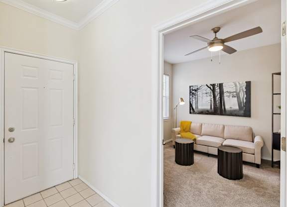 A entryway here at Quail Landing with a bright front door, clean tile flooring, and a view into the living area featuring a beige sofa, yellow throw blanket, neutral carpet, and soft overhead lighting.