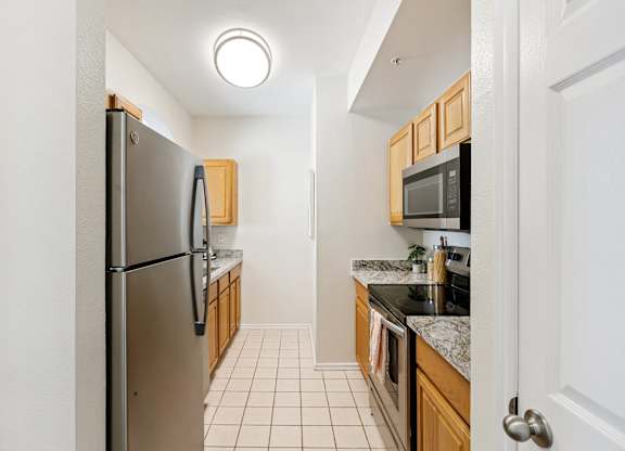 A compact kitchen here at Quail Landing with warm wood cabinets, granite-style countertops, stainless steel refrigerator and range, light tile flooring, and bright overhead lighting in a clean, efficient layout.