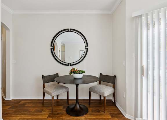 A dining nook here at Quail Landing with warm wood floors, a round dark table, two light upholstered chairs, a simple plant centerpiece, and soft natural light from nearby sliding doors.