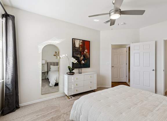 A bedroom here at Quail Landing with soft carpet, a white dresser with gold accents, an arched mirror, abstract wall art, white bedding with a mustard accent pillow, a ceiling fan, and natural light from a window with dark curtains.