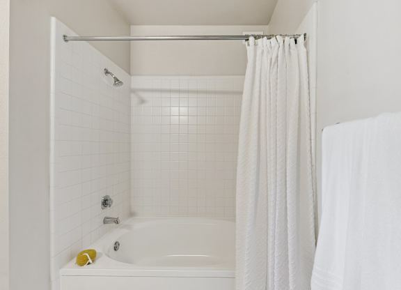 A bathroom shower here at Quail Landing with a white tiled tub surround, curved soaking tub, crisp white shower curtain, chrome fixtures, and a clean, bright, neutral finish.